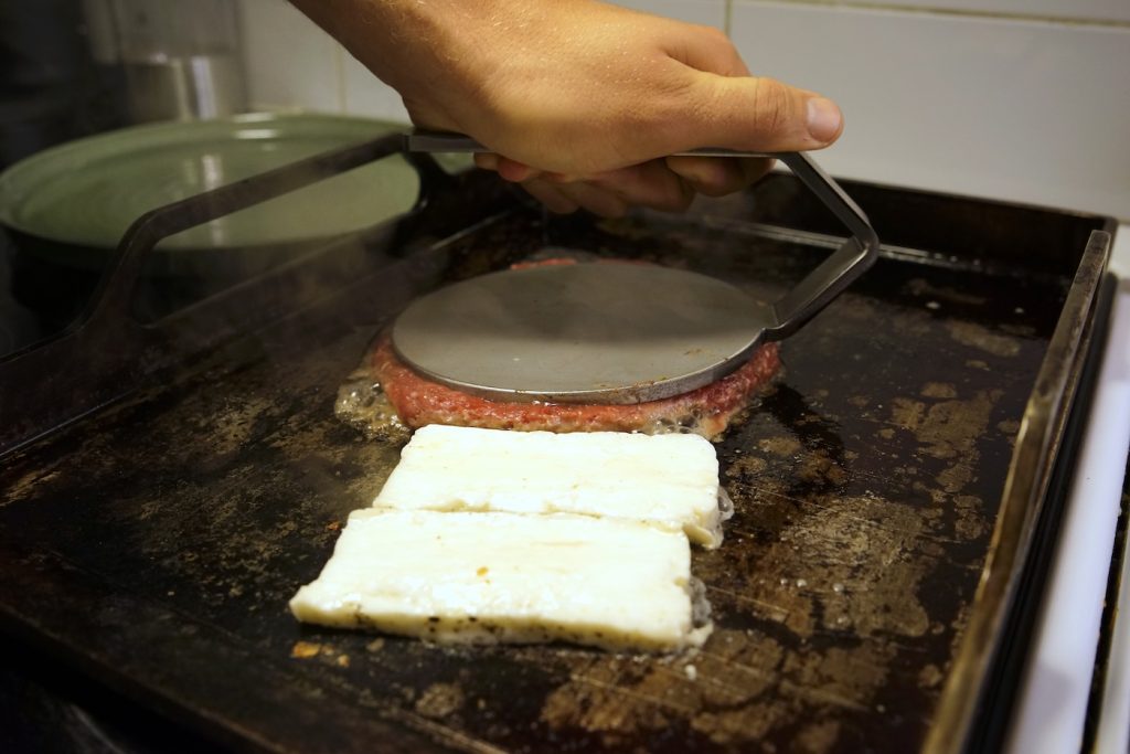 närbild på burgare som smashas och halloumi som steks på tuthult stekbord på induktionshäll